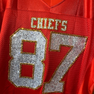 May include: Red football jersey with the word "CHIEFS" and the number "87" in rhinestones.