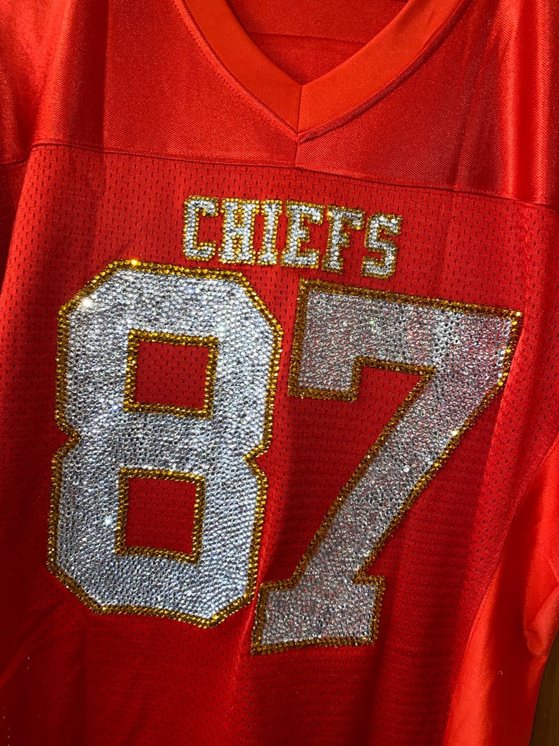 May include: Red football jersey with the word "CHIEFS" and the number "87" in rhinestones.