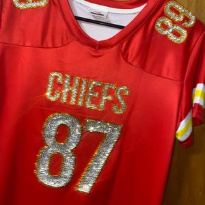 May include: Red football jersey with the word "CHIEFS" and the number "87" in rhinestones. The jersey has gold trim and a white collar.