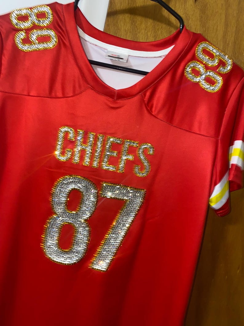 May include: Red football jersey with the word "CHIEFS" and the number "87" in rhinestones. The jersey has gold trim and a white collar.