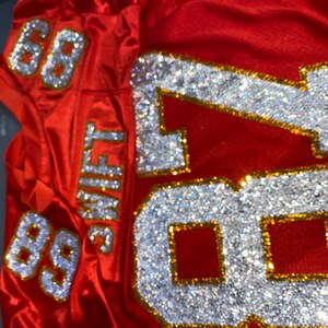 May include: Red football jersey with the number 88 on the sleeves and the number 87 on the front, all decorated with rhinestones. The name "SWIFT" is on the back of the jersey.