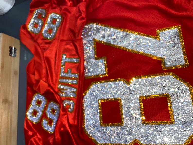 May include: Red football jersey with the number 88 on the sleeves and the number 87 on the front, all decorated with rhinestones. The name "SWIFT" is on the back of the jersey.