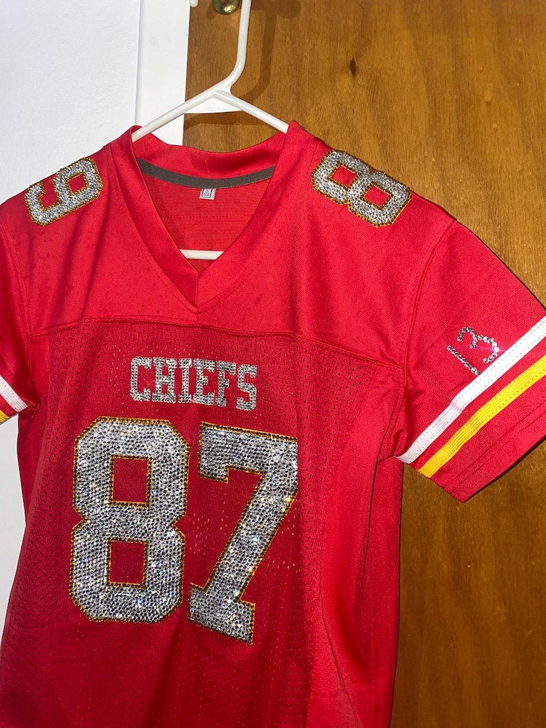 May include: Red football jersey with the number 87 in rhinestones. The jersey has the word "CHIEFS" on the front and the number 8 on each shoulder.