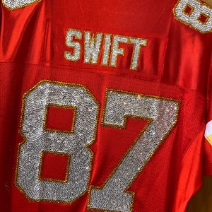 May include: Red football jersey with the number 87 and the name "SWIFT" on the back, all decorated with rhinestones.
