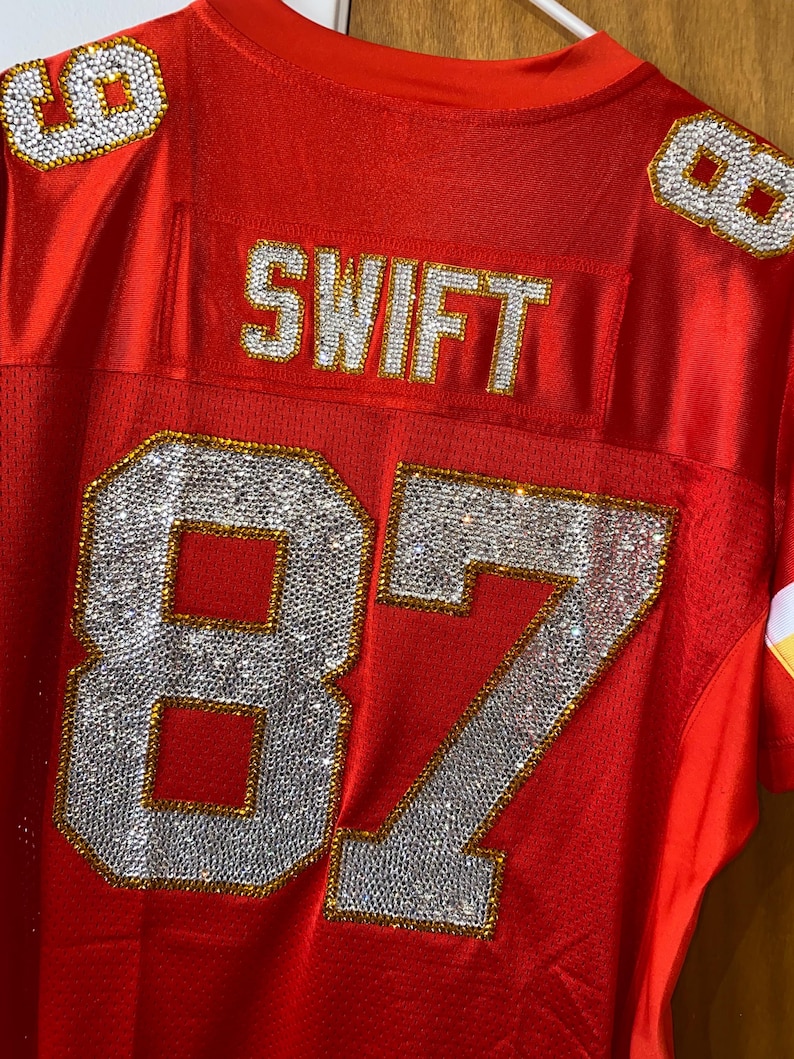 May include: Red football jersey with the number 87 and the name "SWIFT" on the back, all decorated with rhinestones.