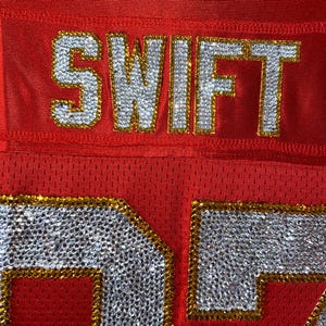 May include: Red football jersey with the name "SWIFT" and the number "27" in rhinestones with gold trim.