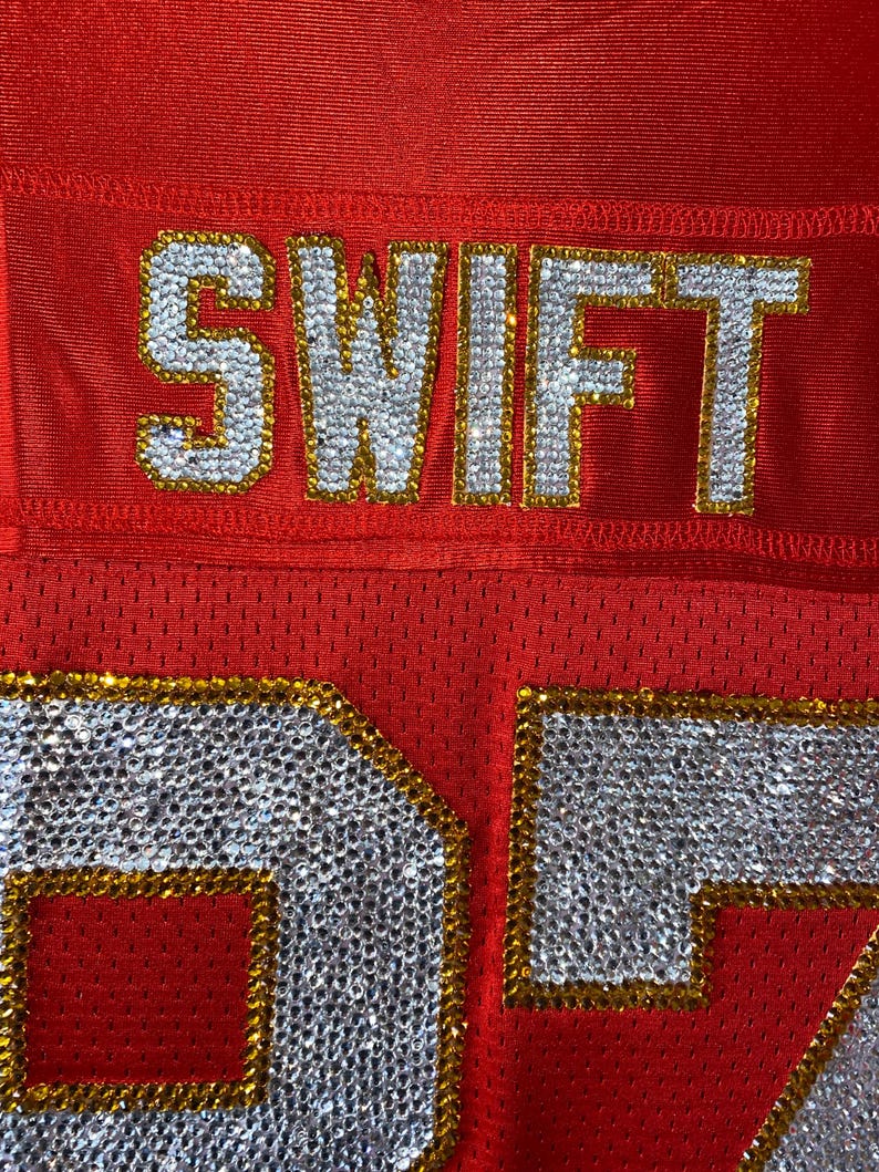May include: Red football jersey with the name "SWIFT" and the number "27" in rhinestones with gold trim.