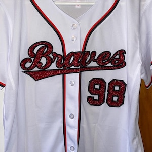 May include: White baseball jersey with red and black trim. The jersey has the word "Braves" in red glitter lettering and the number "98" in red glitter on the front.