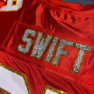 May include: Red football jersey with the number "10" on the shoulder and the word "SWIFT" spelled out in rhinestones on the back.