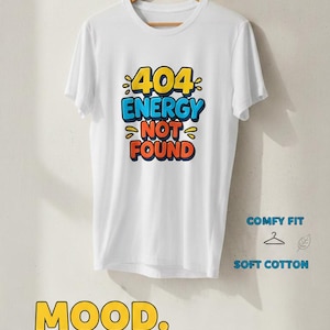 Peut inclure: T-shirt blanc avec l'inscription "404 ENERGY NOT FOUND" en jaune, bleu et rouge. Le t-shirt porte également l'inscription "MOOD. MORNINGS BE LIKE..." en jaune et bleu. Le t-shirt est suspendu à un cintre en bois.