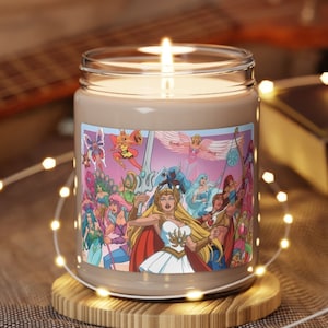May include: A beige scented candle with a She-Ra and the Princesses of Power illustration. The illustration features She-Ra and other characters from the cartoon series. The candle is in a clear glass jar with a brown label. The candle is sitting on a wooden coaster.