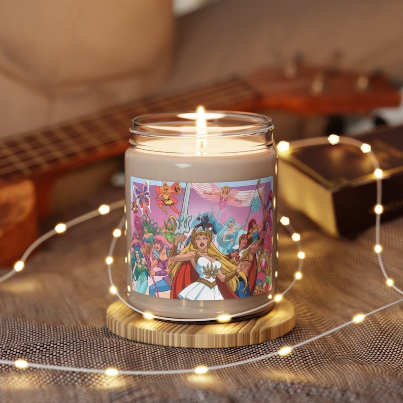 May include: A beige scented candle with a She-Ra and the Princesses of Power illustration. The illustration features She-Ra and other characters from the cartoon series. The candle is in a clear glass jar with a brown label. The candle is sitting on a wooden coaster.