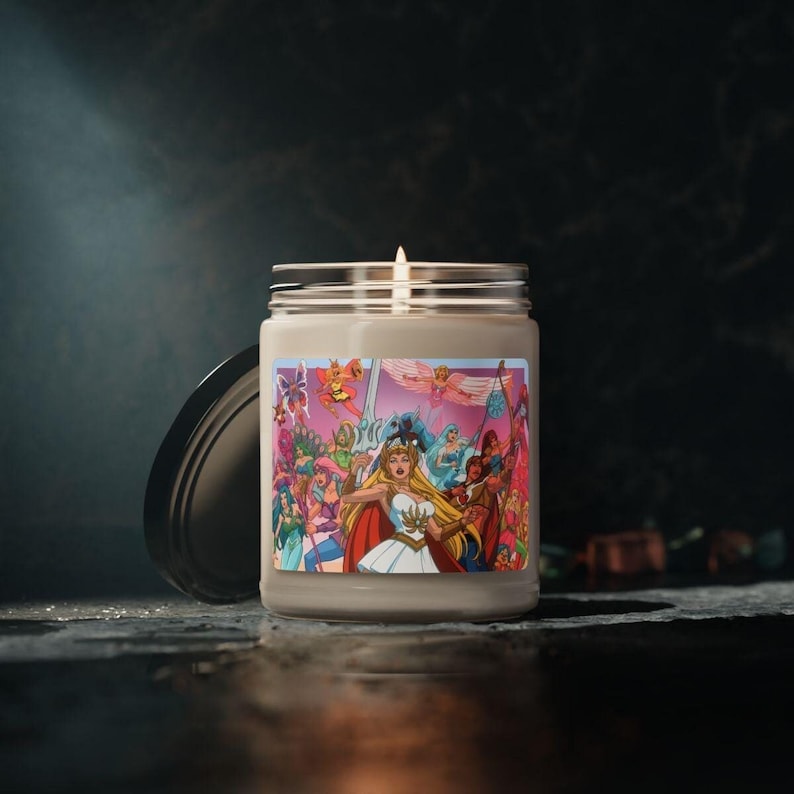 Puede incluir: Un tarro de vidrio para velas con una ilustraci&oacute;n rosa, blanca y azul de un grupo de personajes de dibujos animados. Los personajes son todos mujeres y visten atuendos coloridos. El tarro de vela est&aacute; sobre una superficie oscura.
