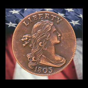 May include: A copper-colored coin with the word "LIBERTY" at the top and the year "1803" at the bottom. The coin features a profile of a woman with flowing hair. The background is an American flag.