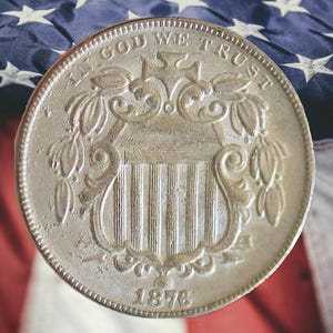 May include: A silver 1876 Shield Nickel coin with the inscription "IN GOD WE TRUST." The coin features a shield design with vertical stripes and is set against a backdrop of the American flag.