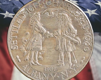 May include: A silver-colored coin with an embossed design of two figures shaking hands, surrounded by the words "SETTLED 1614 CHARTERED 1686 ALBANY N.Y. 1936". The coin is set against a backdrop of the American flag.