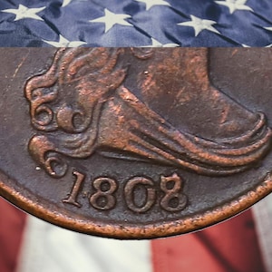 May include: Close-up of a copper coin with the year 1808 visible. The coin features a detailed relief of a figure and is set against a backdrop of the American flag, with its stars and stripes.