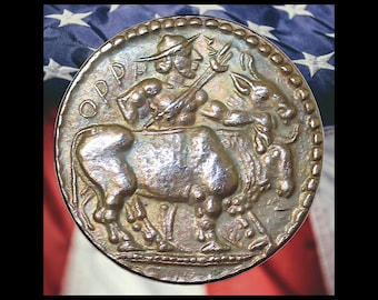 May include: A circular metallic coin featuring a relief of a figure riding a bull. The figure wears a hat and holds a staff. The Greek letters OPH are visible on the left side. The coin has a beaded border and a weathered, dark bronze finish.