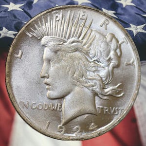 May include: A silver Peace dollar coin from 1924, featuring a profile of Lady Liberty with rays emanating from her crown. The coin is set against a backdrop of the American flag, with red and white stripes and white stars.