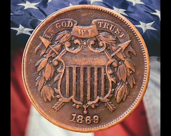 Op de afbeelding: Een koperen twee cent munt uit 1869, met de inscriptie "In God We Trust" en een schildontwerp. De munt is geplaatst tegen een achtergrond van de Amerikaanse vlag, met rode en witte strepen en een blauw veld met witte sterren.