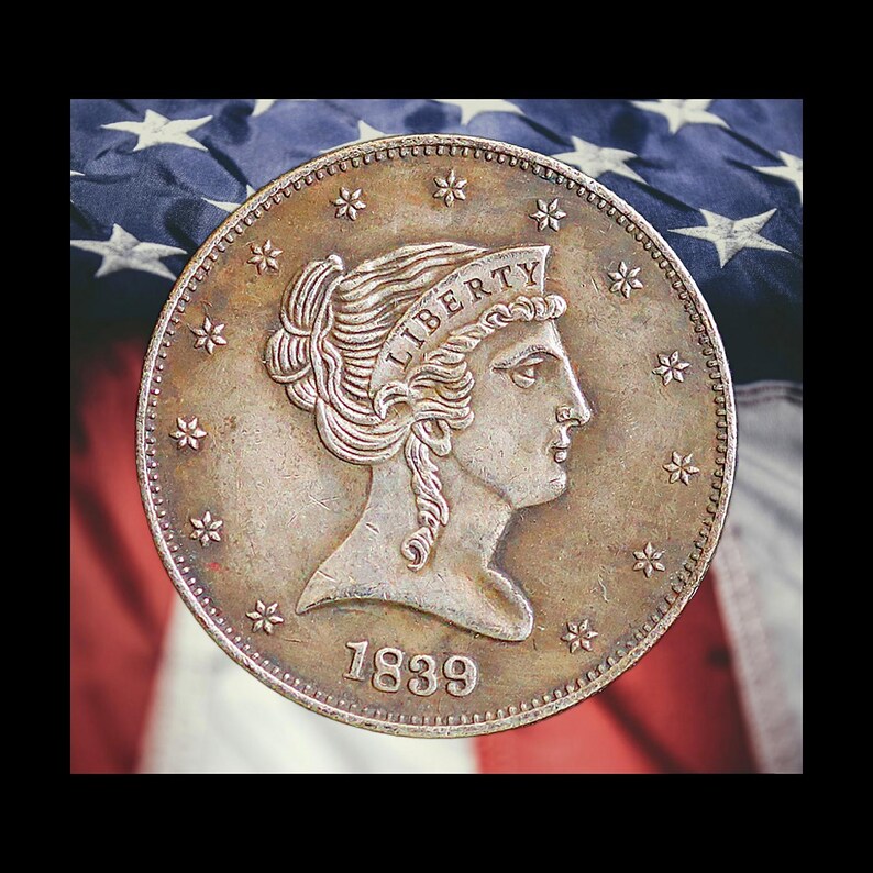 Pu&ograve; includere: Una moneta d'argento con la scritta "LIBERTY" sopra il profilo di una donna. La moneta &egrave; datata 1839 e circondata da stelle. Lo sfondo presenta la bandiera americana. Il diametro &egrave; di circa 3,8 cm.