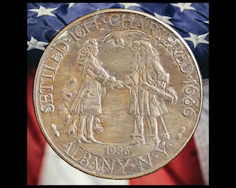 May include: A round bronze-toned commemorative coin featuring two figures shaking hands in the center. The text reads SETTLED 1614 CHARTERED 1686 and ALBANY N.Y. 1936. An eagle is depicted above the figures, with pine trees visible on the left side.