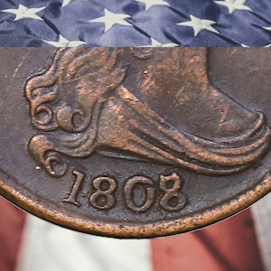 May include: A close-up of a copper-colored coin featuring a profile relief of a figure wearing a cap, with the year 1808 embossed clearly at the bottom. The coin has a weathered, metallic texture and is set against a blurred American flag background.