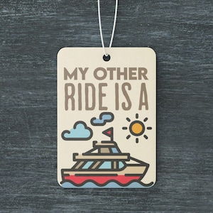 Pode incluir: Um aromatizador de carro bege com um cordão branco para pendurar. Apresenta uma ilustração de desenho animado de um barco com uma bandeira vermelha, branca e azul, um céu azul com nuvens brancas e um sol amarelo. O texto "MY OTHER RIDE IS A" está impresso em marrom no aromatizador.