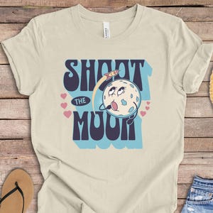 Puede incluir: Camiseta de color natural con la frase "SHOOT THE MOON" en una fuente retro. El diseño incluye una luna de dibujos animados con un cohete y corazones. La camiseta se muestra sobre una superficie de madera con accesorios.