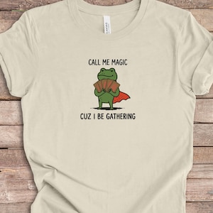 Puede incluir: Una camiseta de color natural con un gráfico de una rana verde que lleva una capa roja y sostiene un abanico de cartas de juego. El texto sobre la rana dice CALL ME MAGIC y debajo de la rana dice CUZ I BE GATHERING en fuente negra.