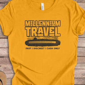 Könnte beinhalten: Ein senfgelbes Kurzarm-T-Shirt mit einem schwarzen Grafikdruck, der MILLENNIUM TRAVEL liest, einer Abbildung eines Raumschiffs und dem Text FAST | DISCREET | CASH ONLY, der darunter in einer fetten, serifenlosen Schrift gedruckt ist.
