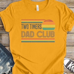 Könnte beinhalten: Ein senfgelbes Kurzarm-T-Shirt mit einem Grafikdruck aus horizontalen Streifen in Rot, Orange, Gelb und Blaugrün. Der Text "TWO TIMERS DAD CLUB" ist in einer dunkelblauen serifenlosen Schrift gehalten, ergänzt durch eine halbkreisförmige Sonne.