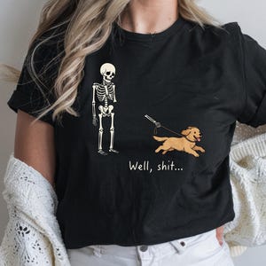 May include: A black short-sleeved t-shirt featuring a graphic of a white skeleton holding a leash attached to a running golden-coloured dog. Below the graphic, the text reads "Well, shit..." in a simple white font. The shirt has a crew neckline.