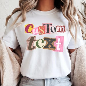 Op de afbeelding: Wit T-shirt met de woorden "Custom Text" in een collage-stijl, met behulp van verschillende kleuren en patronen. Het woord "WHITE" staat onderaan het shirt.