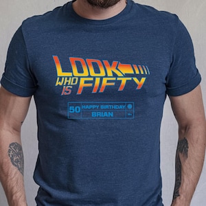 May include: Heather navy blue t-shirt with the text "LOOK WHO IS FIFTY" in a retro design. The shirt also includes "50 HAPPY BIRTHDAY BRIAN" in a blue box. A casual tee for a milestone birthday.