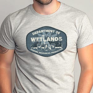 May include: Athletic heather gray t-shirt featuring a blue and white graphic. The graphic reads "DEPARTMENT OF WETLANDS" with illustrations of birds and the text "KEEPING RESOURCES PRODUCTIVE". The shirt is made of a soft, comfortable material.