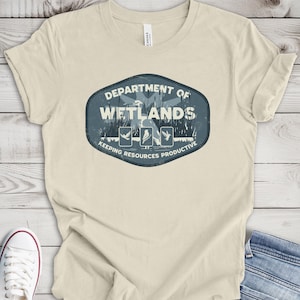 May include: A natural-colored t-shirt with a blue and white graphic that reads "Department of Wetlands" with illustrations of birds and the text "Keeping Resources Productive." The shirt is paired with blue jeans and white sneakers.