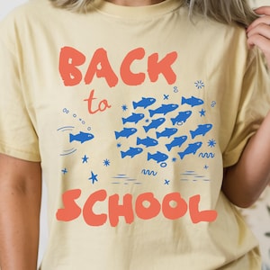Può includere: Una t-shirt a maniche corte giallo pallido con il testo rosso BACK TO SCHOOL. Tra le parole c'è la grafica di un grande banco di pesci blu che nuotano insieme, accompagnata da piccole icone di stelle e onde blu, con la scritta BUTTER sotto.