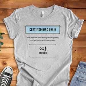 以下が含まれることがあります： 青い枠の中に「CERTIFIED BIRD BRAIN」という文字が書かれた薄い灰色のTシャツ。枠の下には「Birds obsessed with creating habitats, gaining food, laying eggs, and drawing cards.」という文章が書かれている。その下には、鳥が描かれた無限大記号と「PER BIRD」という文字が書かれている。Tシャツの下部には「ATHLETIC HEATHER」という文字が書かれている。