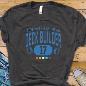 May include: Dark gray heather t-shirt with the text "SEVENTEEN & CERTIFIED DECK BUILDER 17" in blue. The shirt features a design with playing card graphics and a series of colored dots. The words "DARK GREY HEATHER" are printed at the bottom.