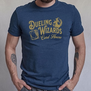 May include: Navy heather t-shirt with gold text that reads "Dueling Wizards Card House" and a graphic of a wizard and cards. The shirt is short-sleeved.