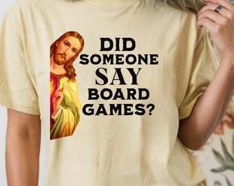 May include: A butter-coloured short-sleeve t-shirt featuring a graphic of Jesus on the left side and the black text "DID SOMEONE SAY BOARD GAMES?" printed on the right. The shirt has a crew neckline and a relaxed fit, showcasing a clear, bold design.