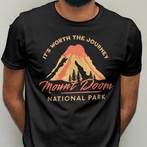 May include: Black t-shirt featuring a mountain graphic with the text "It's Worth the Journey" and "Mount Doom National Park" in a vintage style. The graphic includes a volcano and trees in shades of orange and yellow.