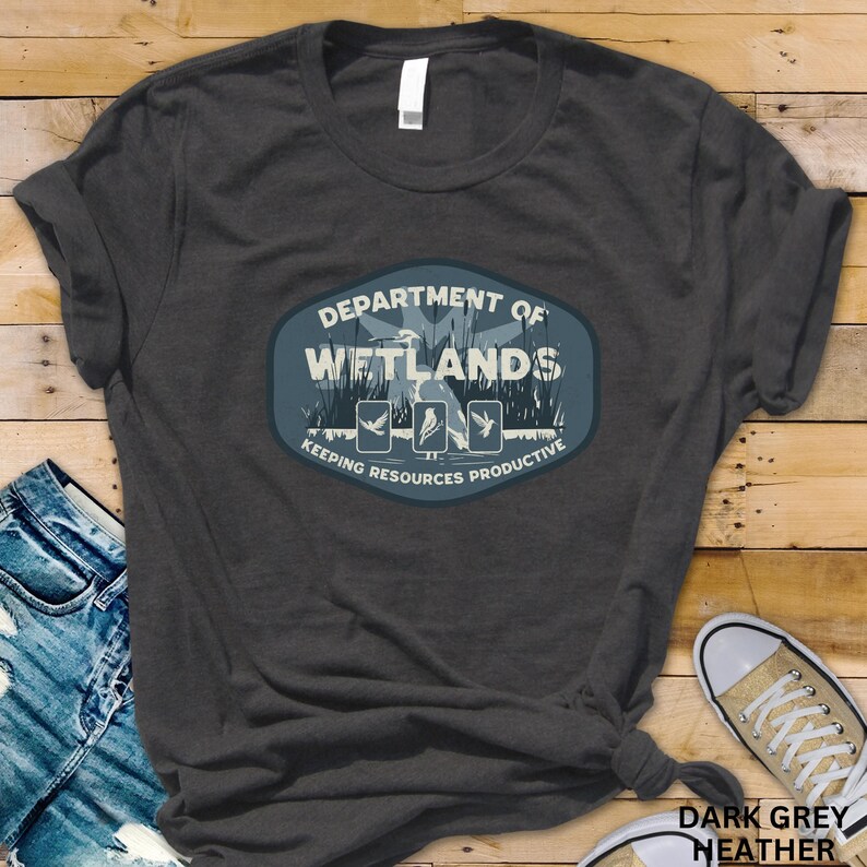 May include: Dark grey heather t-shirt featuring a graphic with the text "DEPARTMENT OF WETLANDS" and the phrase "KEEPING RESOURCES PRODUCTIVE". The design includes illustrations of birds and plants. The shirt is a casual wear item.