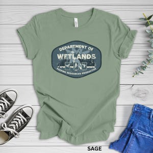 May include: Sage green t-shirt featuring a graphic that reads "Department of Wetlands" with illustrations of birds and the text "Keeping Resources Productive." The shirt is paired with black sneakers and blue jeans.