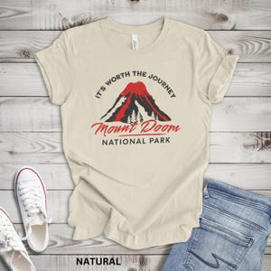 May include: Cream-colored t-shirt with the text "It's Worth the Journey" above a graphic of a red and black mountain with the words "Mount Doom National Park" below. The word "NATURAL" is printed at the bottom.