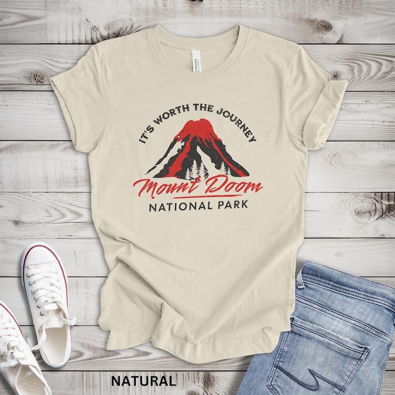 May include: Cream-colored t-shirt with the text "It's Worth the Journey" above a graphic of a red and black mountain with the words "Mount Doom National Park" below. The word "NATURAL" is printed at the bottom.