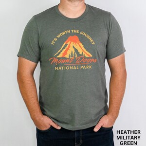 May include: Heather military green t-shirt with a graphic of a mountain and the text "It's Worth the Journey" and "Mount Doom National Park". The shirt is a crew neck and has short sleeves.
