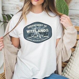 May include: White t-shirt featuring a distressed oval design with the text "DEPARTMENT OF WETLANDS" and the phrase "KEEPING RESOURCES PRODUCTIVE." The design includes illustrations of birds and plants. The shirt is made of a soft material.
