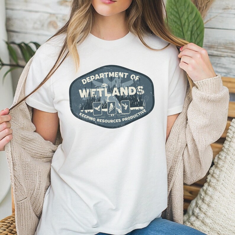 May include: White t-shirt featuring a distressed oval design with the text "DEPARTMENT OF WETLANDS" and the phrase "KEEPING RESOURCES PRODUCTIVE." The design includes illustrations of birds and plants. The shirt is made of a soft material.